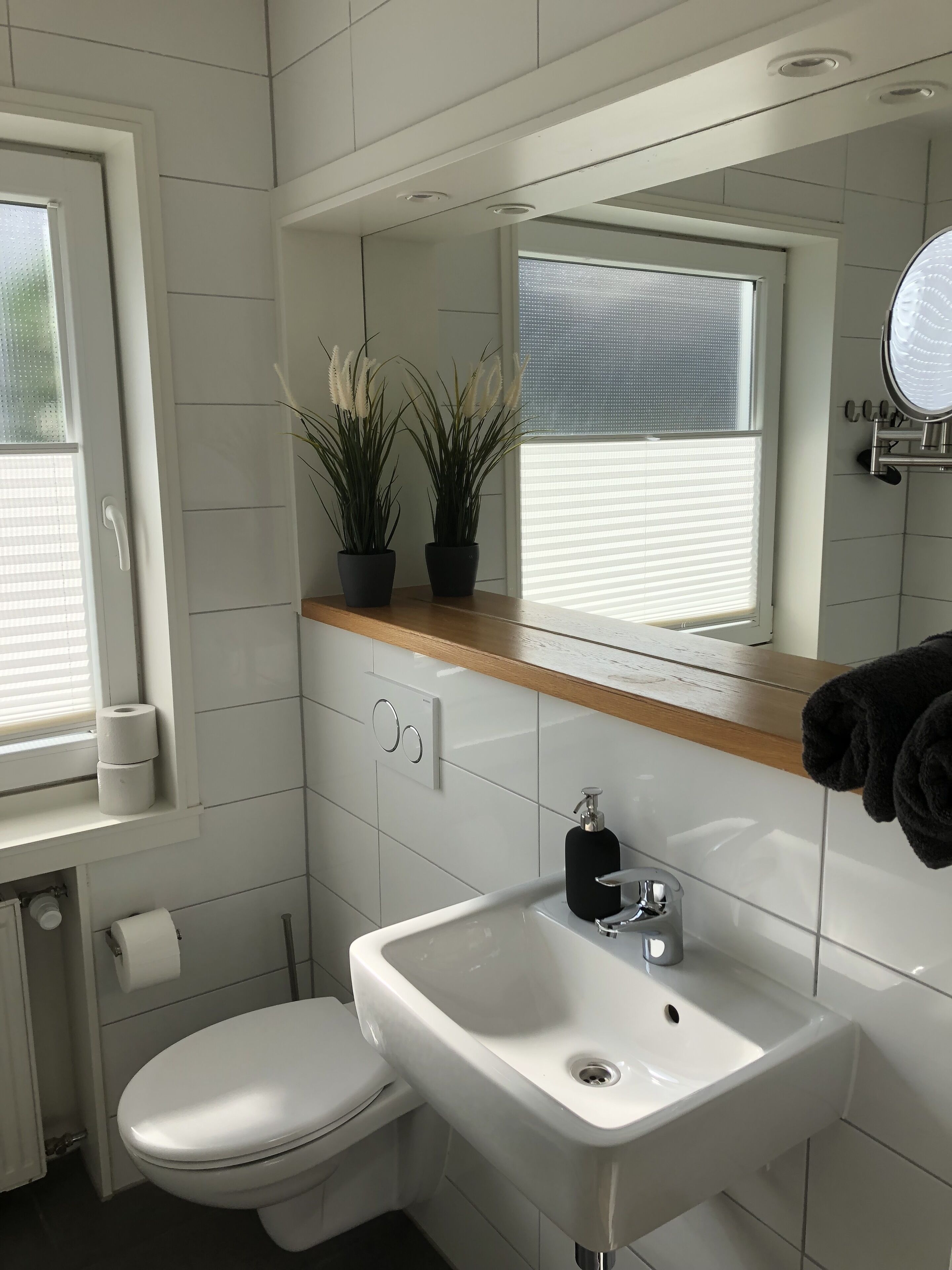 A modern bathroom with a white toilet, a sink with a black soap dispenser, and two potted plants on a wooden shelf above the sink.