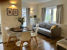 Una sala moderna con un sofá gris, sillas blancas, una mesa de comedor redonda con individuales tejidos y un florero con flores.