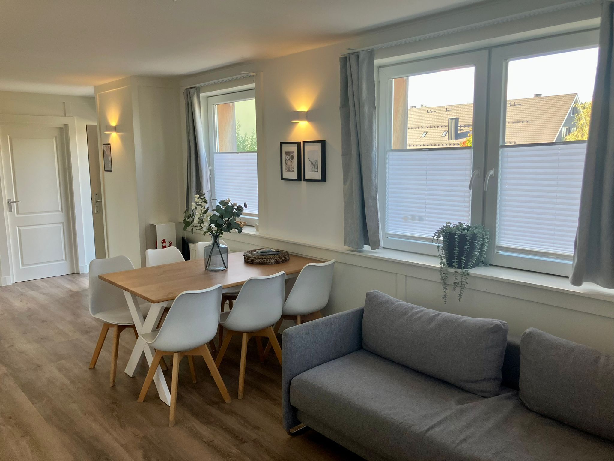A modern living room with a wooden dining table, white chairs, a grey sofa, and large windows with curtains.