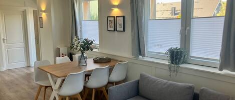 A modern living room with a wooden dining table, white chairs, a grey sofa, and large windows with curtains.