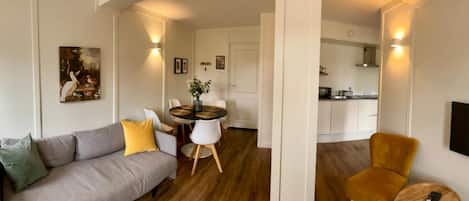 A modern living room with a grey sofa, yellow and green cushions, a dining table with chairs, a kitchen area with a TV, and a wooden floor.