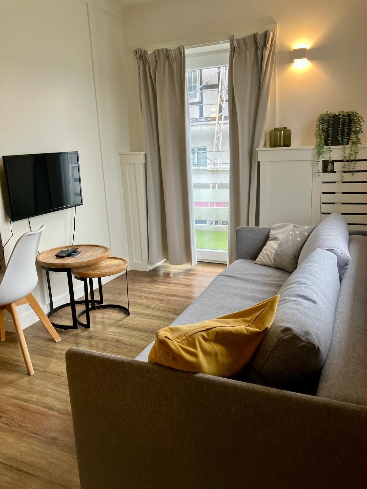 Een moderne woonkamer met een grijze bank, een houten salontafel, een witte fauteuil, een flatscreen televisie en een schuifdeur naar een buitenruimte.