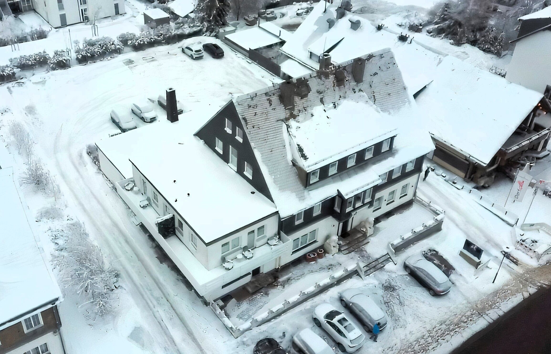 A snow-covered residential area with multiple houses, a driveway, and parked cars.