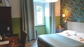 A hotel room with a bed, a desk, a chair, a window with curtains, and a wall with a floral pattern.
