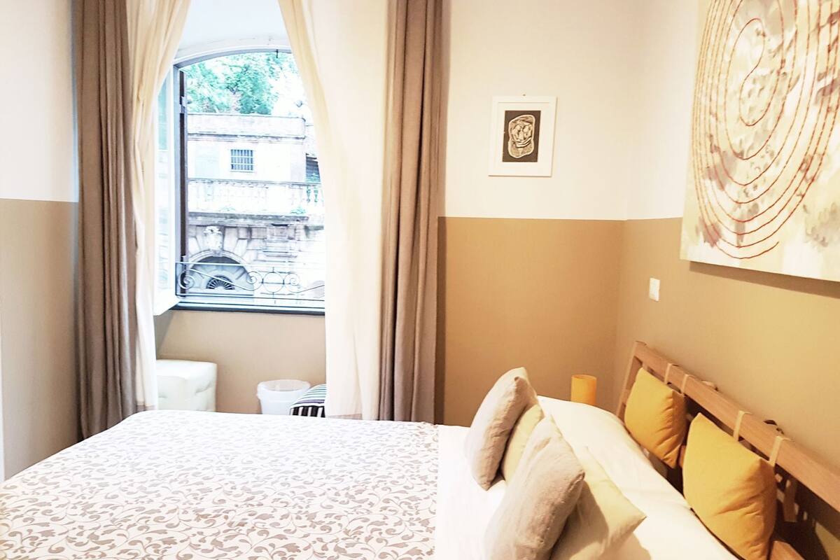 Une chambre d’hôtel avec un lit, des rideaux, une fenêtre donnant sur un immeuble, une petite table de chevet et une œuvre d’art accrochée au mur.