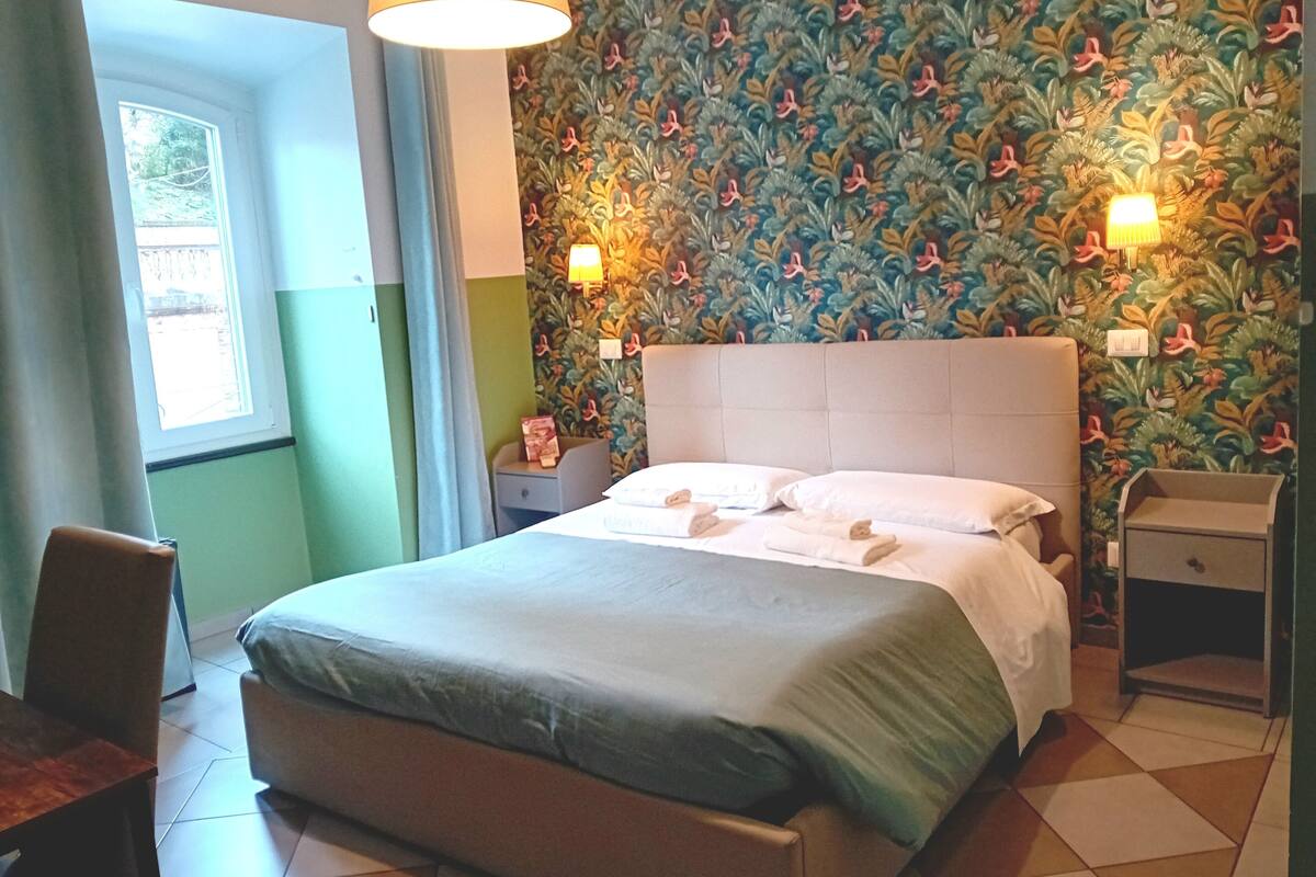Un lit bien fait, avec des oreillers et une table de chevet, une fenêtre avec des rideaux et un mur orné d’un motif floral.