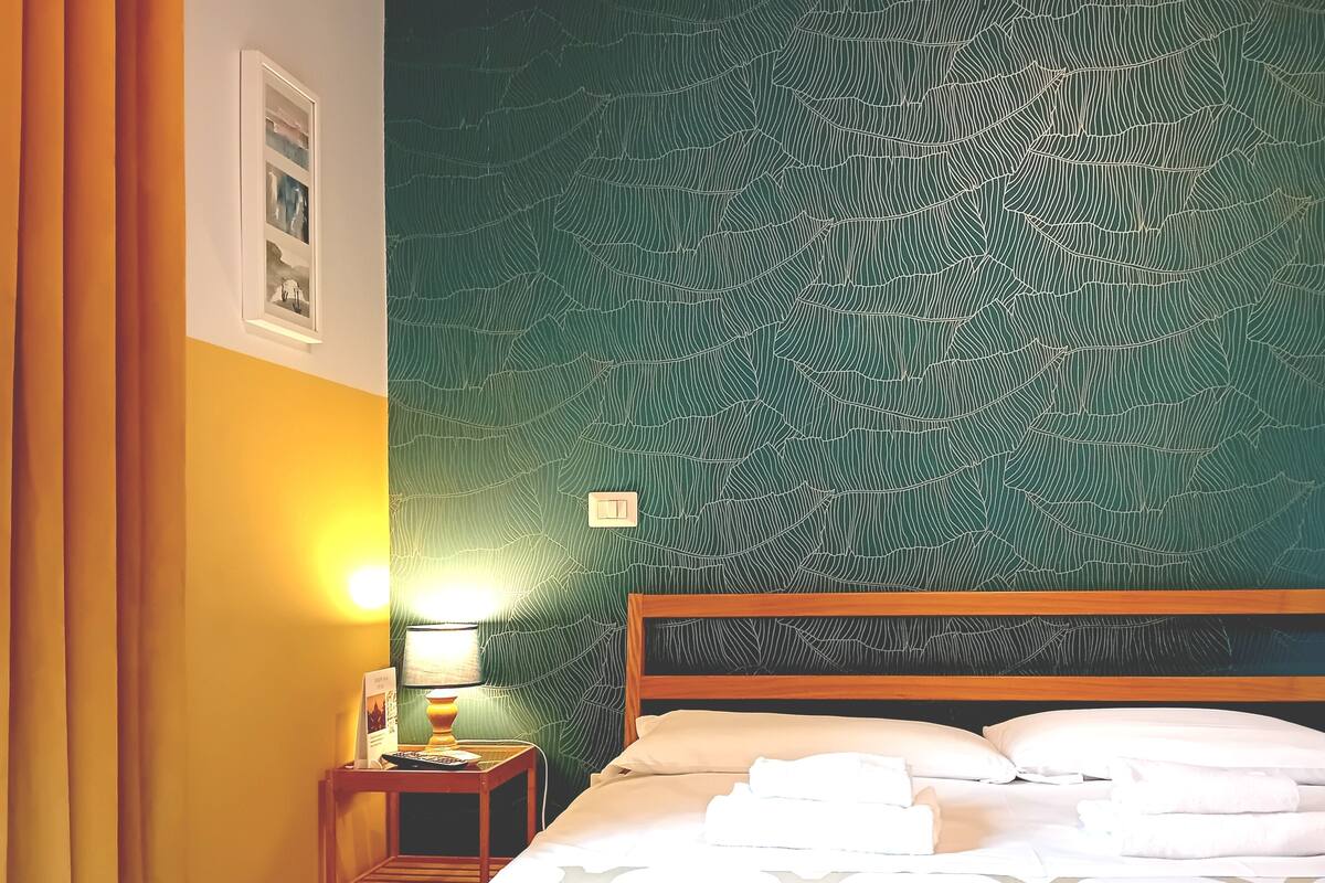 Une chambre d’hôtel avec un mur texturé de couleur verte, une tête de lit en bois et un lit recouvert de draps et de serviettes blancs.