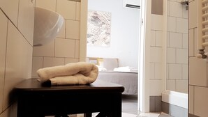 Un baño de hotel con una toalla sobre un taburete, una zona de ducha y una cama visible a través de una puerta abierta.