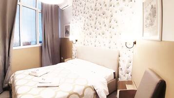 Una camera d'albergo con un letto, una sedia, una finestra con tende e pareti caratterizzate da un motivo decorativo, illuminata da un punto luce a parete.