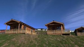 Two wooden cabins with balconies, situated on a grassy hill with a clear blue sky.
