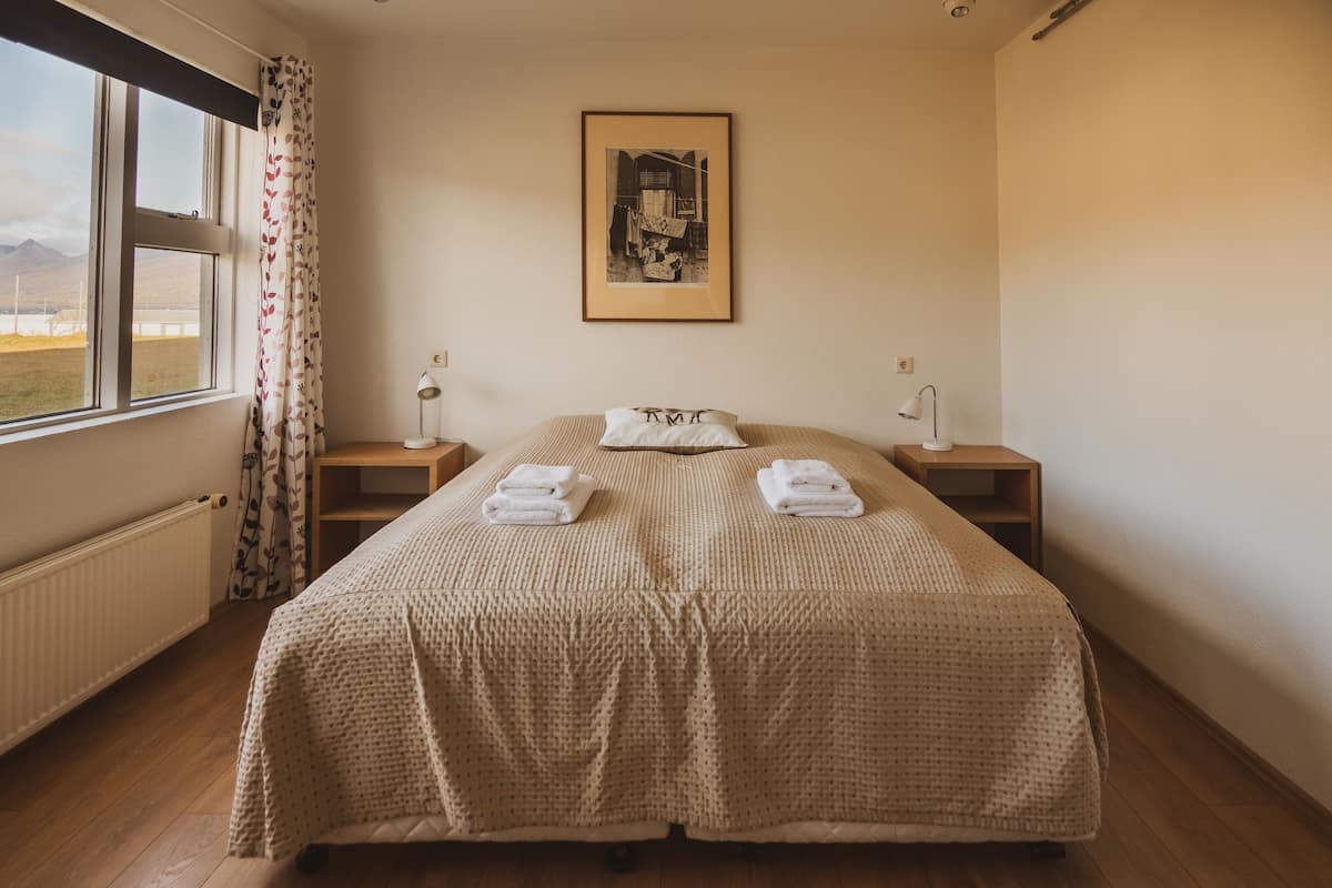 Ein Doppelbett mit einer beigen Steude, zwei Nachttische mit Lampen, ein gerahmtes Bild an der Wand und ein Fenster mit Bergblick.