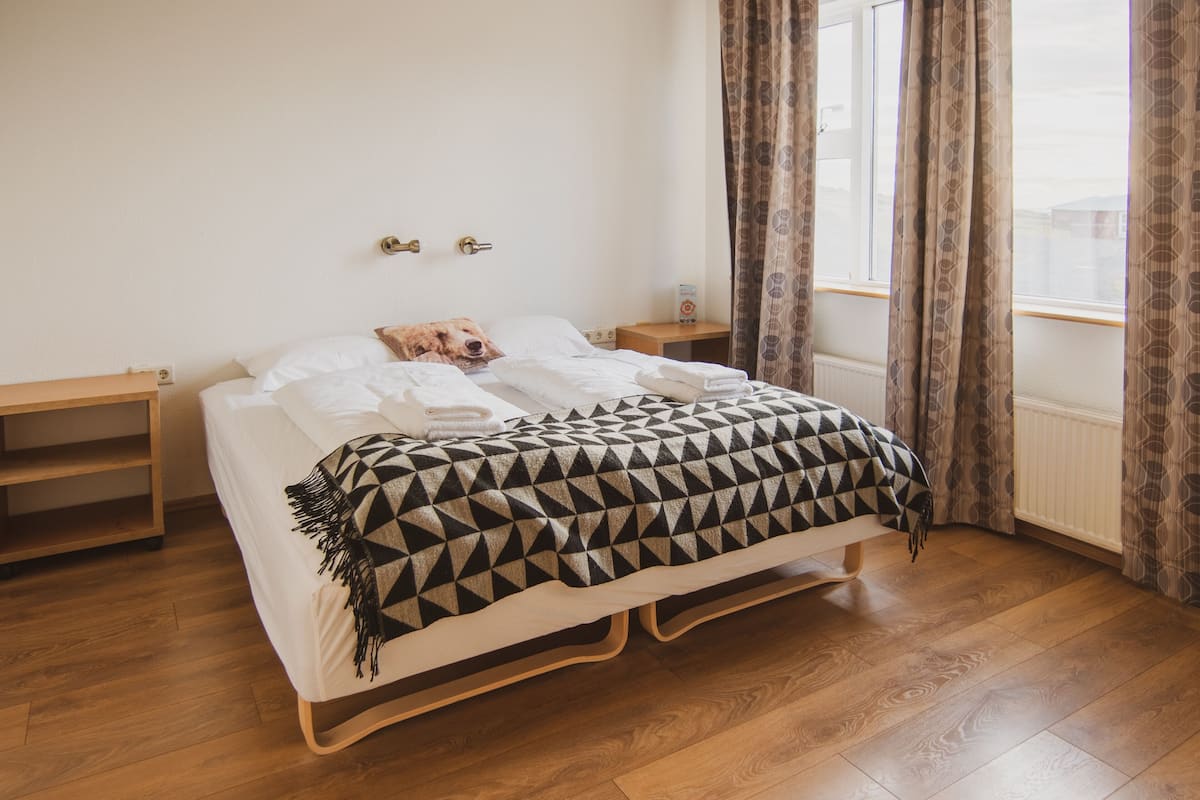 Ein ordentlich bezogenes Bett mit einer geometrisch gemusterten Decke, Nachttische aus Holz und einem Fenster mit Vorhängen.