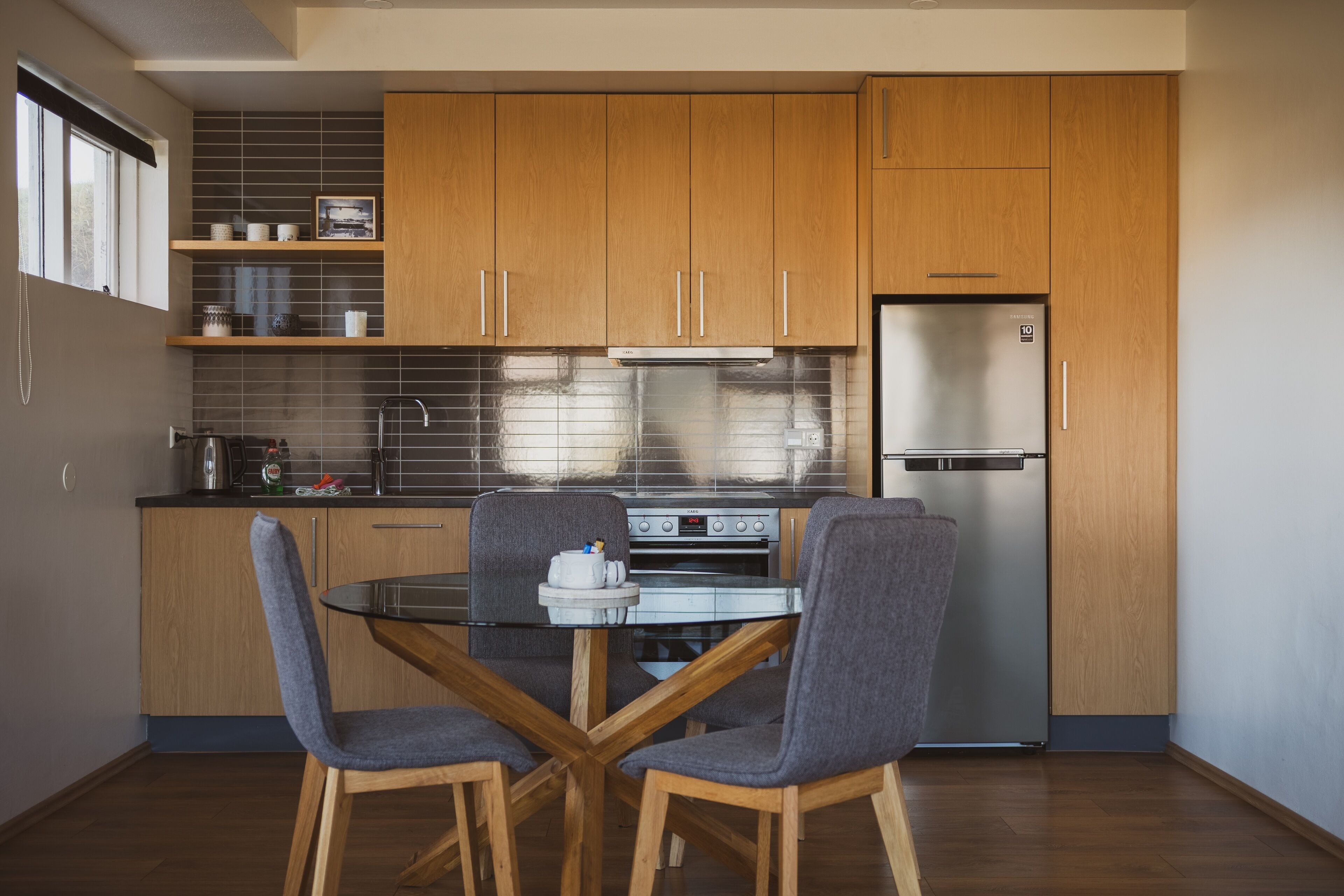 A modern kitchen with wooden cabinets, a stainless steel refrigerator, and a glass dining table with chairs.