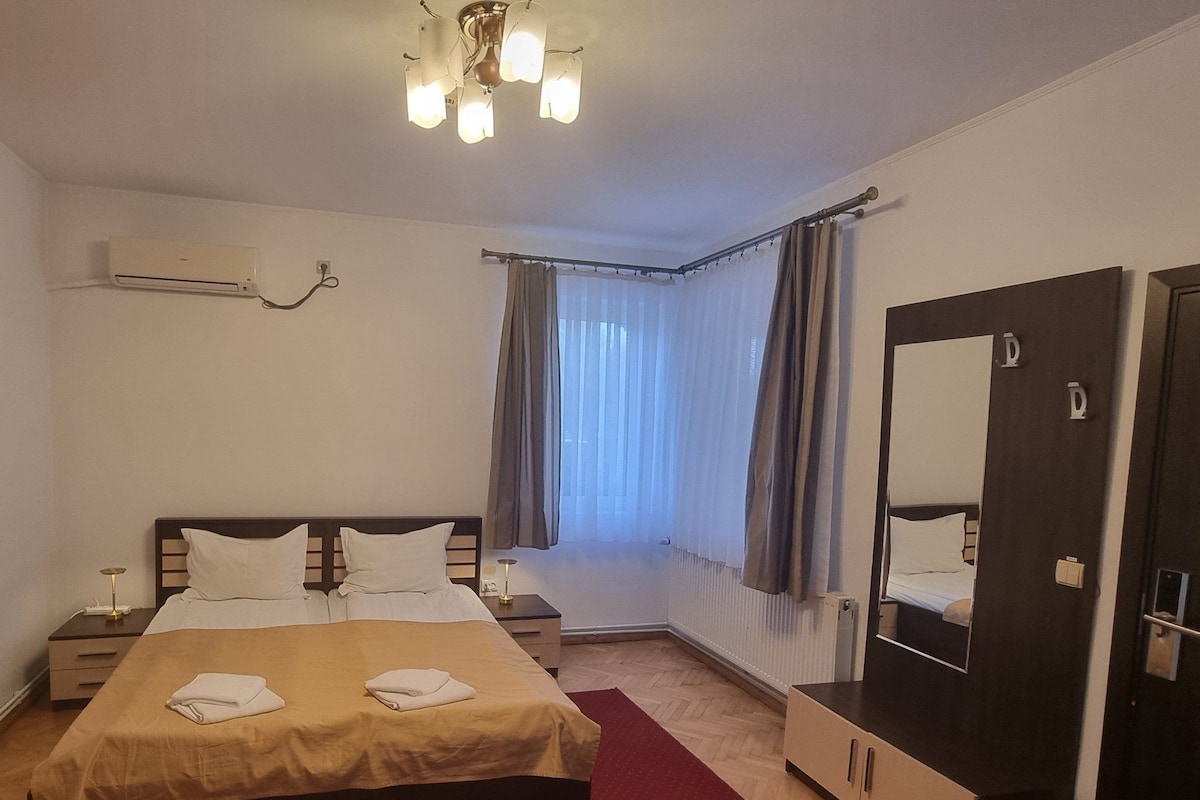 A hotel room with a bed, bedside tables, a mirror, an air conditioning unit, and a window with curtains.