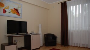 Una sala de estar con televisor, un pequeño aparador, una silla, un tapete rojo y una ventana con cortinas.