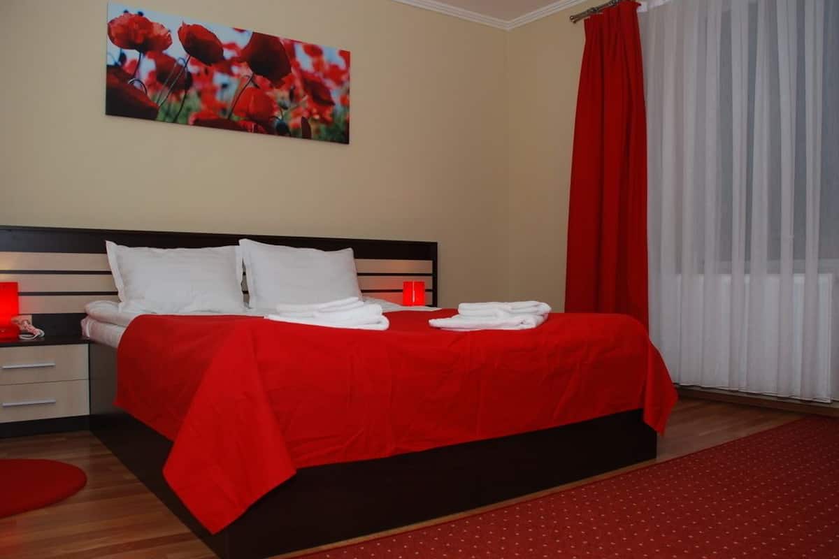 Un lit bien fait, avec du linge de lit blanc et une couverture rouge, une table de chevet avec une lampe rouge et un tableau représentant des fleurs rouges accroché au mur.