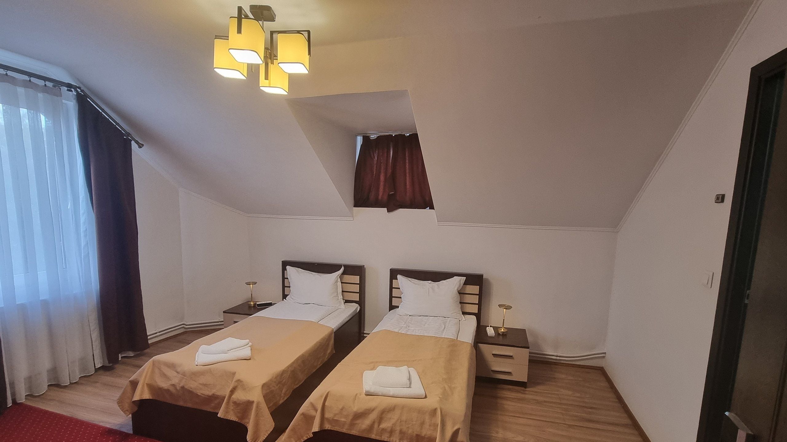 Una habitación pequeña y bien iluminada con dos camas, una mesita de noche y una ventana con cortinas.
