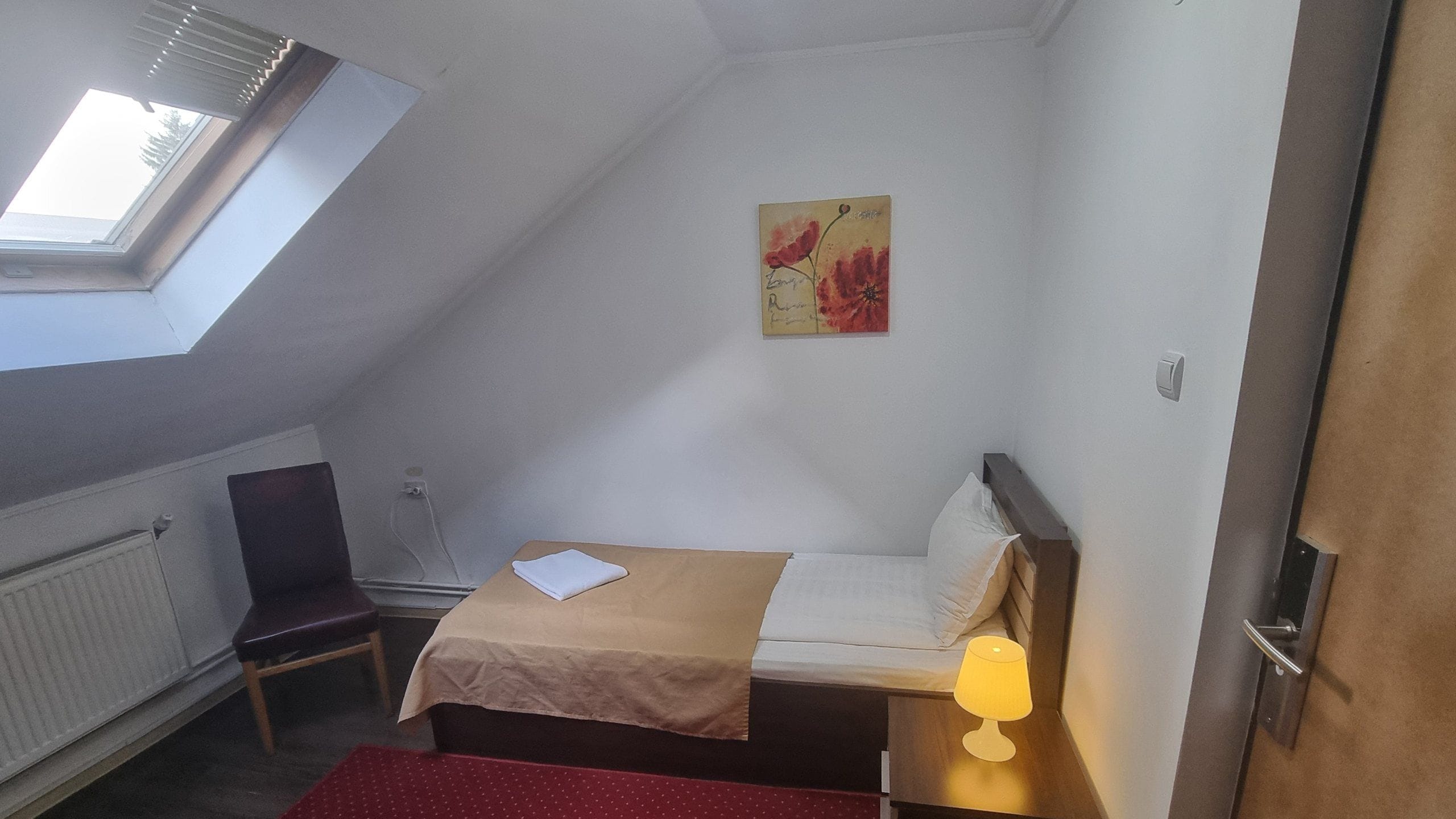 A small, compact room with a bed, a chair, a radiator, and a painting on the wall.
