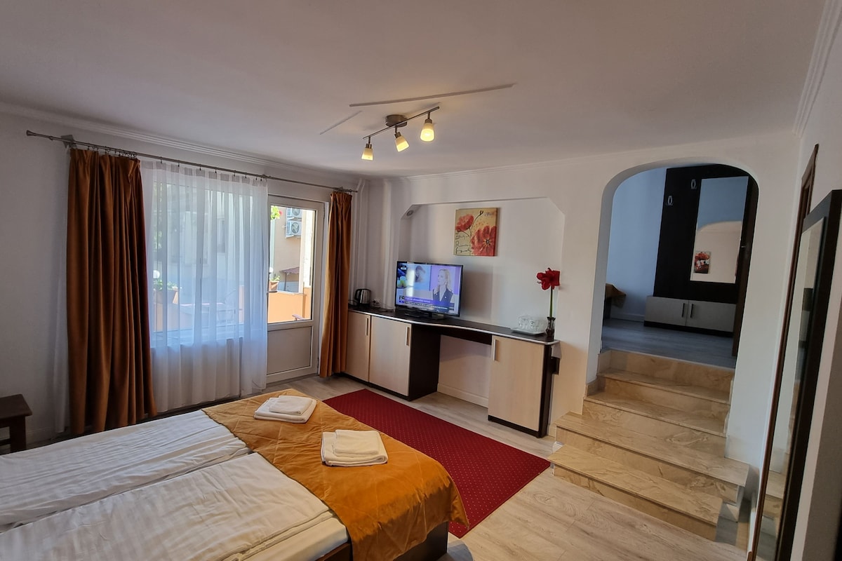 A hotel room with a bed, a desk with a television, a red rug, and a view of the outside.