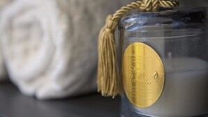 A clear glass candle with a golden label and a rope handle, placed next to a rolled white towel. - Suite Vogue Visconti (Voghera)
