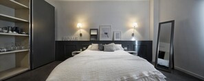 A modern hotel room with a large bed, bedside tables, a wardrobe, and a built-in shelf with kitchenware. - Suite Vogue Visconti (Voghera)