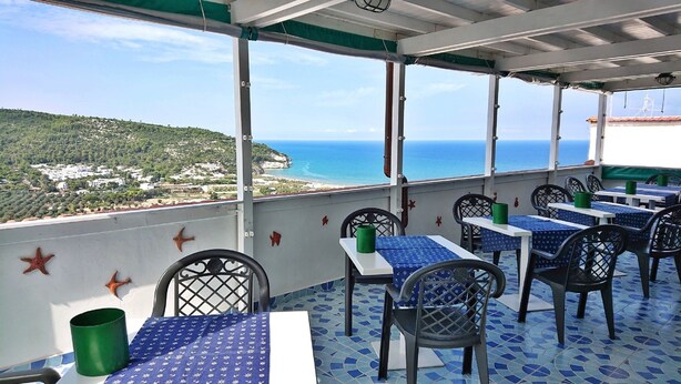 Eine Terrasse mit Tischen und Stühlen, die auf eine Küstenlandschaft mit Häusern und einem klaren Himmel blickt.