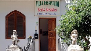 Eine Villa mit einem Schild, auf dem 'Villa Passiatuo Bed & Breakfast' und eine Telefonnummer stehen. Vor dem Eingang führen Treppen hinauf, daneben befindet sich ein Briefkasten und auf jeder Seite ein Standbild.