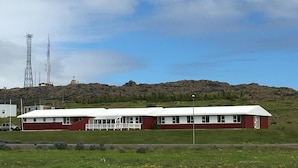 A long, single-story building with a white roof and red walls, situated on a grassy hill with a rocky background. - Helgafell Hostel (Djupivogur)