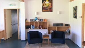 A room with wooden ceiling beams, two blue armchairs, a small table, and bookshelves filled with books and decorative items. - Helgafell Hostel (Djupivogur)