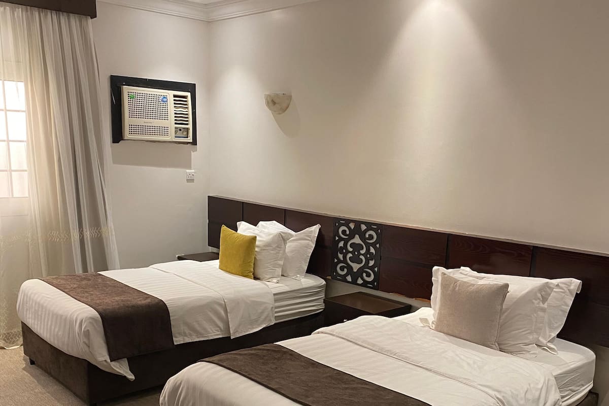 Une chambre d’hôtel avec deux lits, une table de chevet, un climatiseur fixé au mur et une fenêtre avec des rideaux.