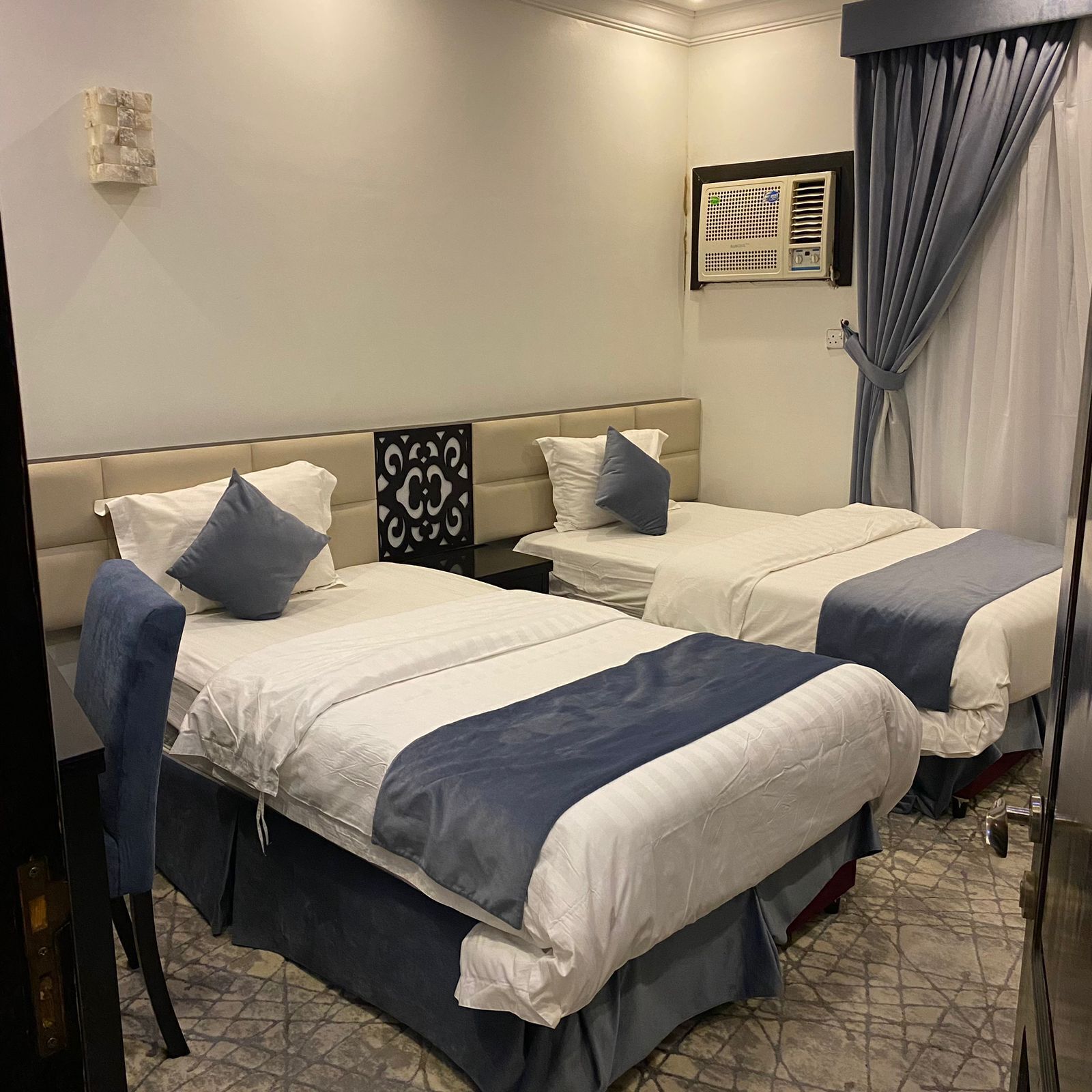 Habitación de hotel con dos camas, cada una con cubrecama azul y blanco, cabecera con panel decorativo, mesita de noche y unidad de aire acondicionado fijada a la pared.
