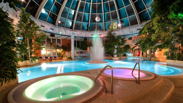 A large indoor swimming pool with multiple hot tubs, a waterfall feature, and a glass-domed ceiling. - Vital Hotel Westfalen Therme (Bad Lippspringe)
