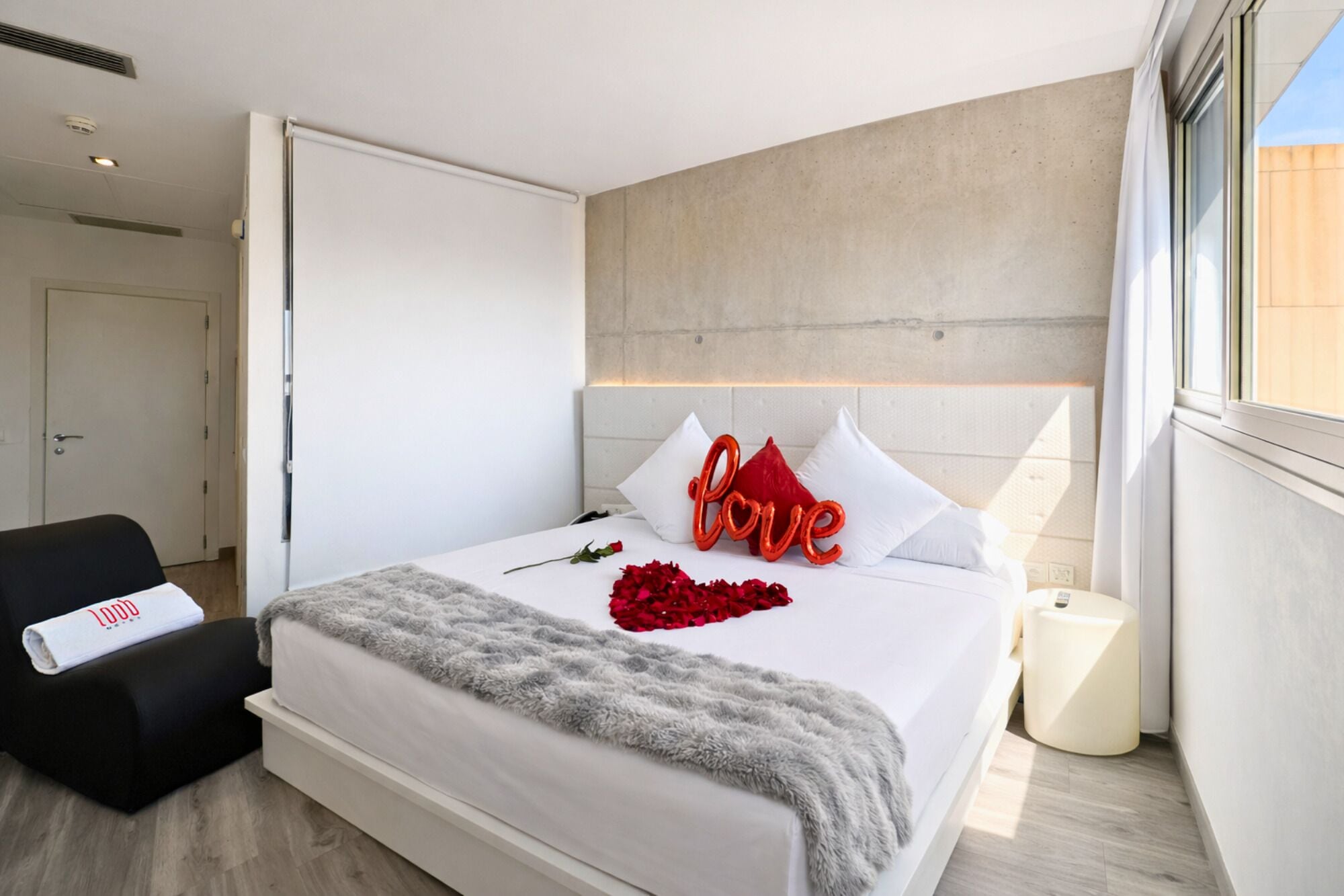 Una habitación de hotel moderna con una cama, una silla negra, una mesita de noche y un ventanal grande.