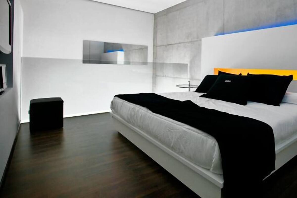 A modern hotel room with a large bed, a small bedside table, a wall-mounted phone, and a dark wooden floor.