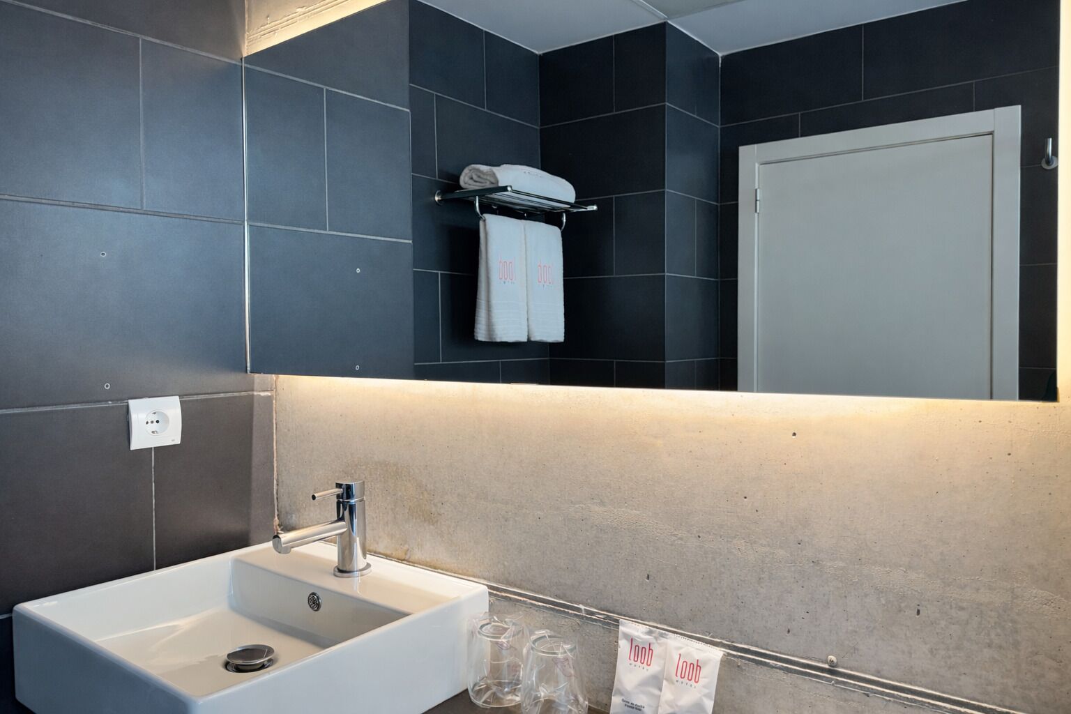 Un baño moderno con un lavamanos blanco, paredes de azulejos oscuros, un perchero y un espejo.