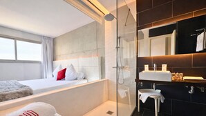 Una habitación de hotel moderna con una cama grande, un box de ducha con paredes de vidrio, un tocador con espejo y vistas al exterior.