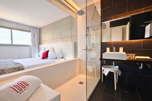 Una habitación de hotel moderna con una cama grande, un box de ducha con paredes de vidrio, un tocador con espejo y vistas al exterior.