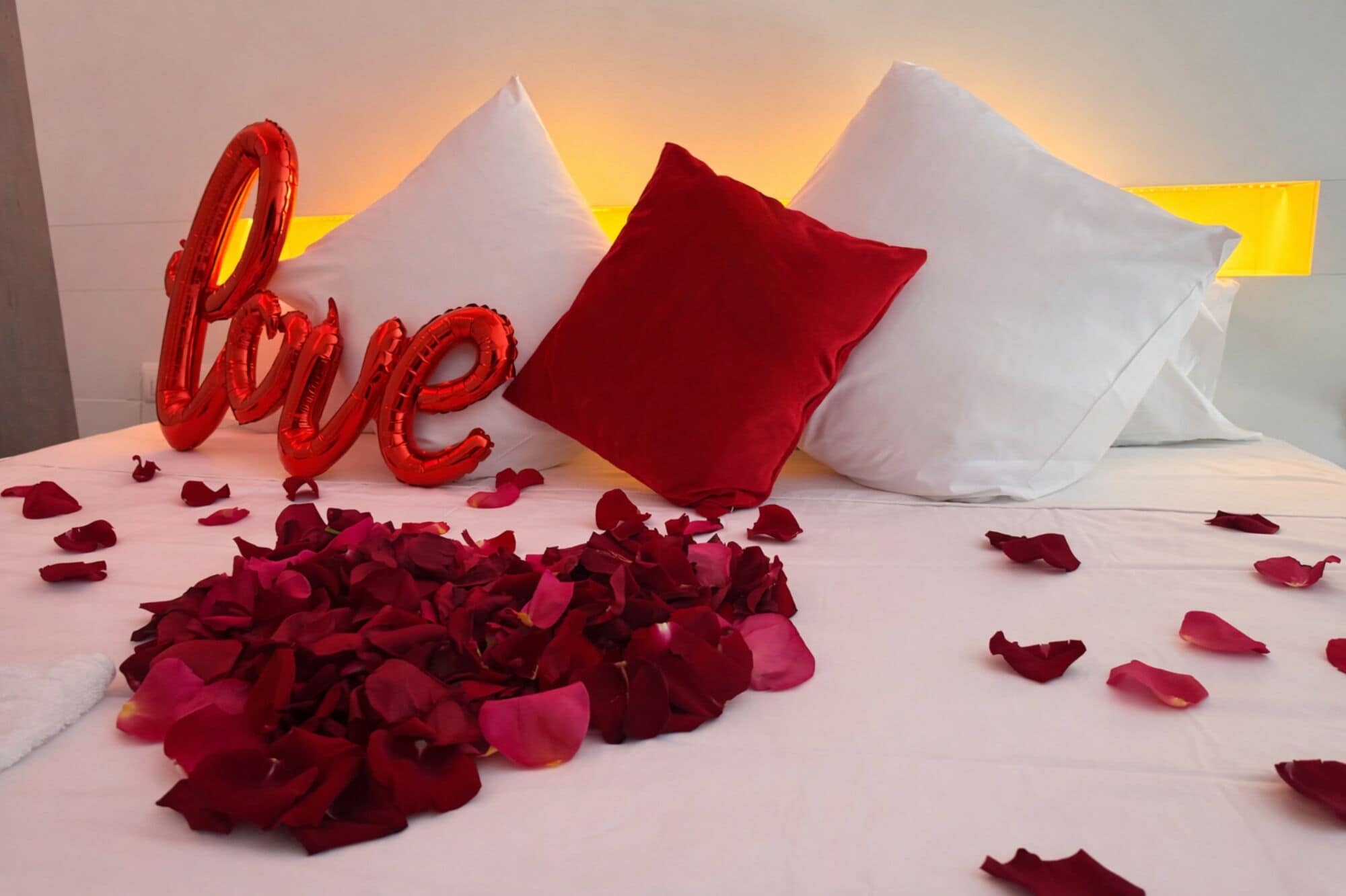 Un lit parsemé de pétales de rose, avec des oreillers rouges et blancs, et un ballon rouge sur lequel est écrit le mot « amour ».