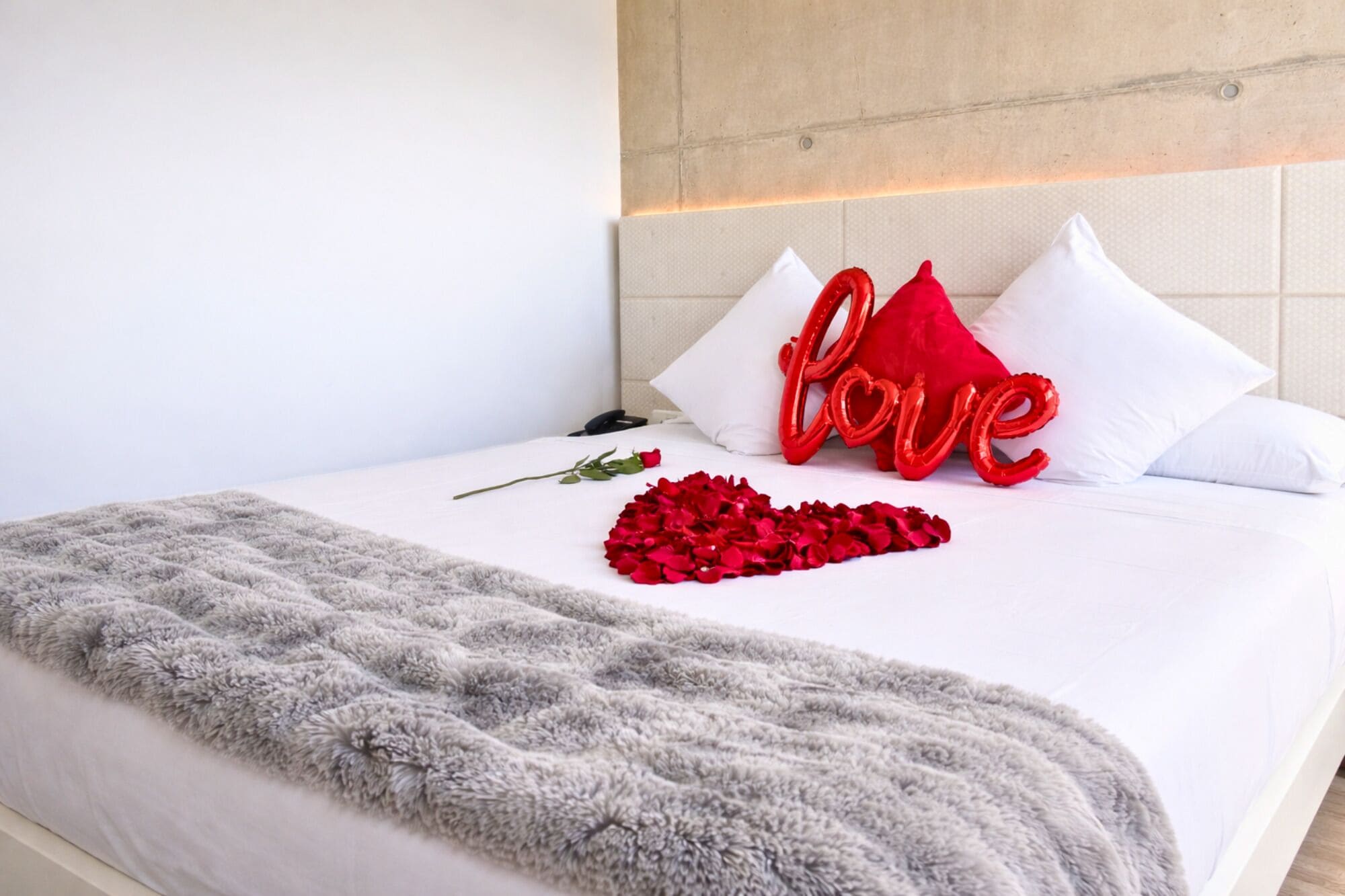 A neatly made bed with a fluffy gray blanket, a single rose, and red heart-shaped decorations on the pillows.