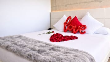 A neatly made bed with a fluffy gray blanket, a single rose, and red heart-shaped decorations on the pillows.