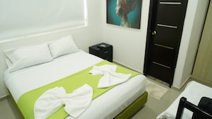 A neatly made bed with white linens and a green accent, a small bedside table with a phone, a door with a handle, and a wall-mounted artwork. - Hotel Sun River (Barranquilla)