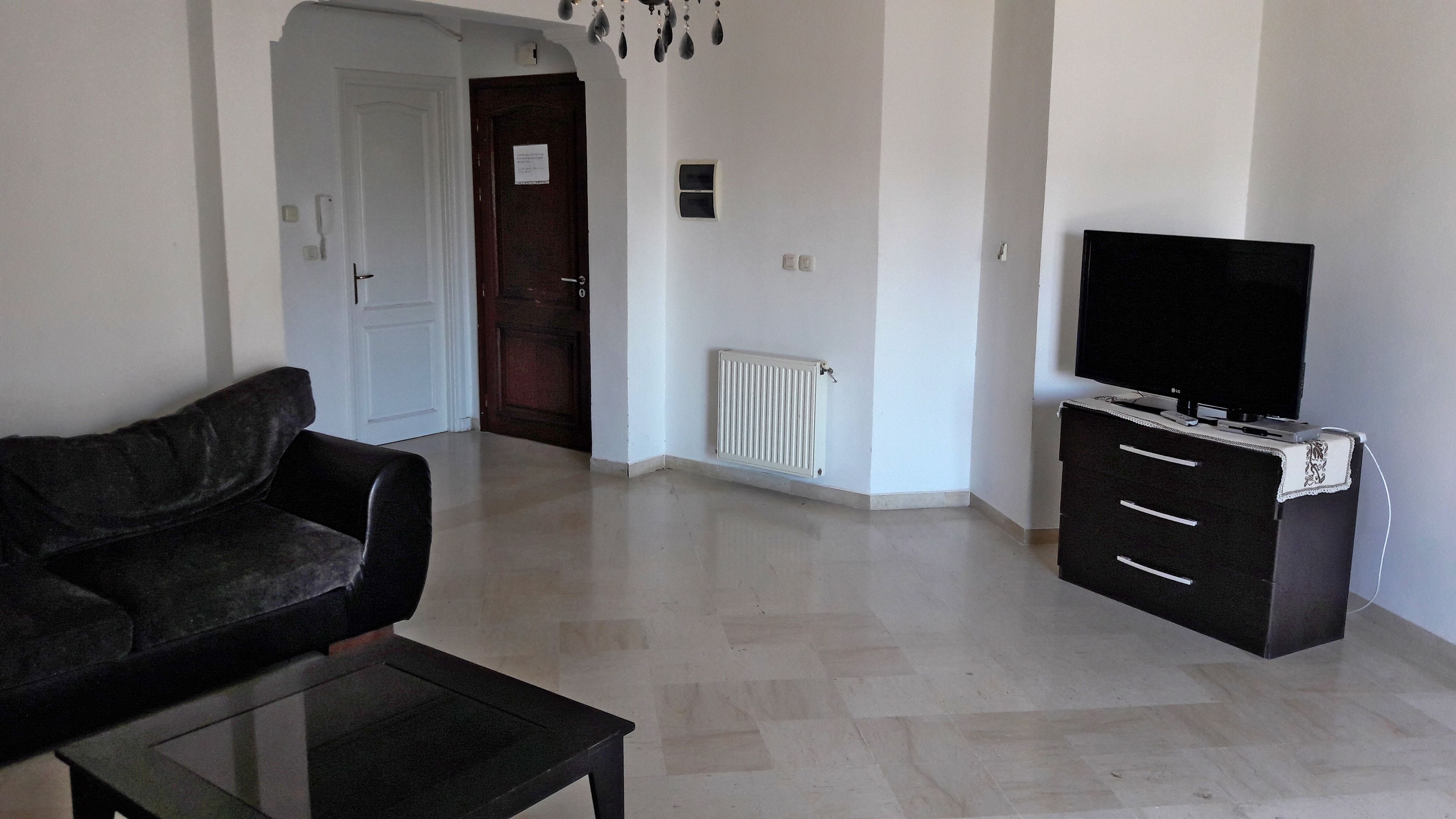 A living room with a sofa, a coffee table, a TV on a cabinet, a radiator, and a door.