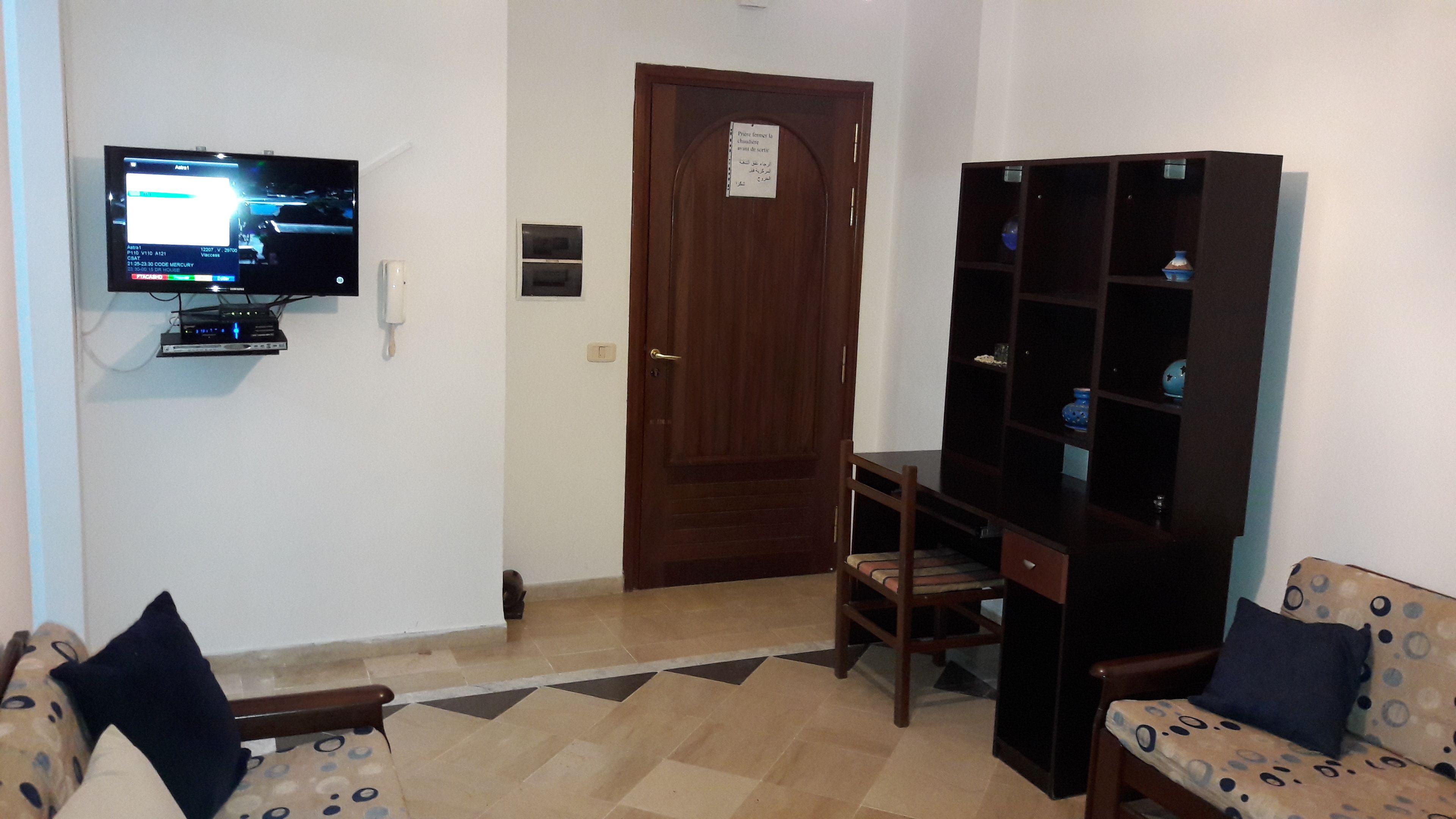 A living room with a television, a wooden door, a bookshelf, and a patterned sofa.