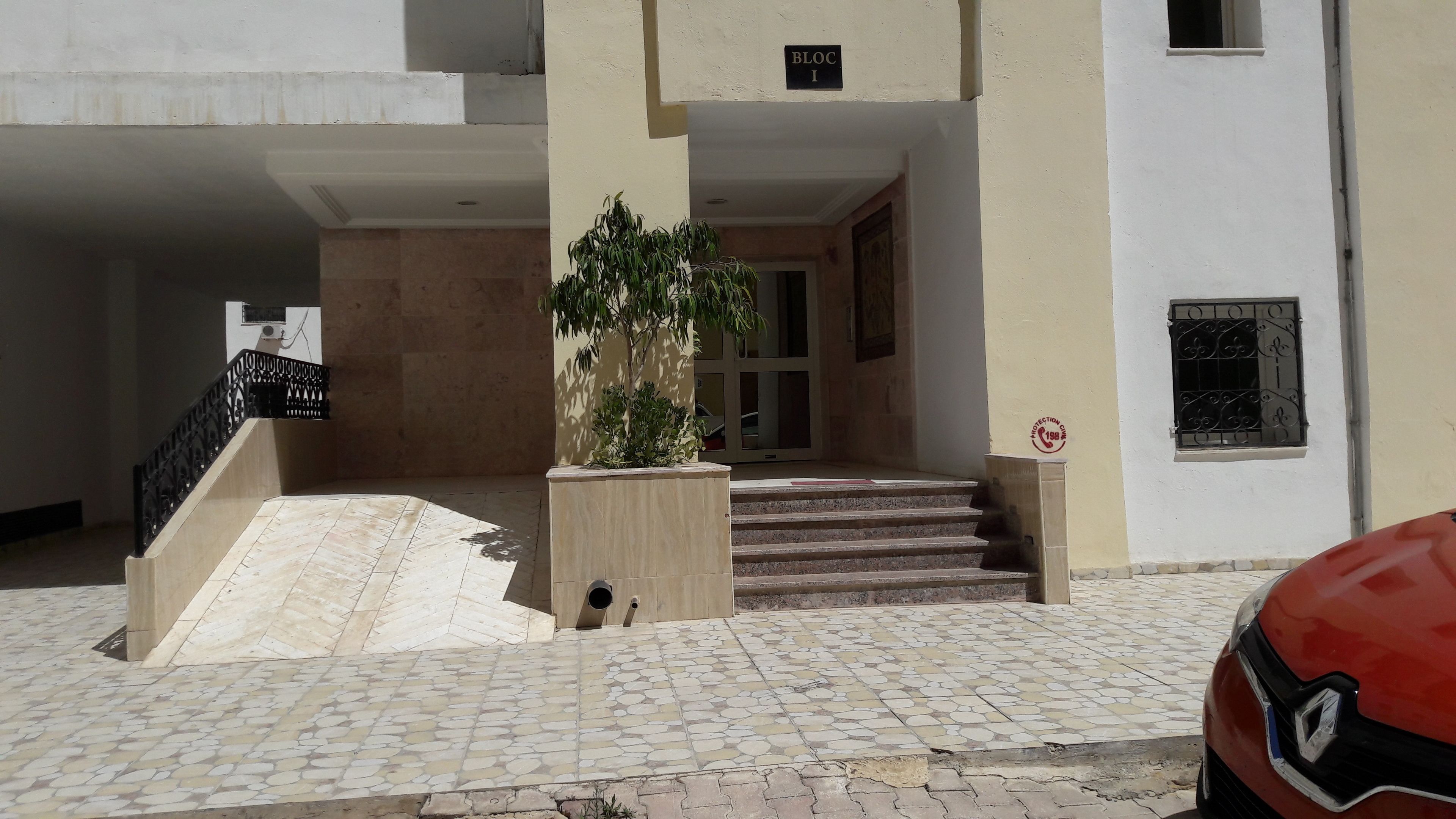 A building entrance with a tiled staircase leading to a door, a metal railing, and a sign indicating 'BLOC I'.