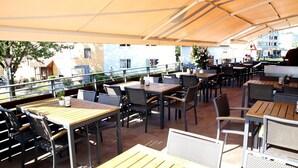 Un café en plein air avec des tables et des chaises disposées sous une marquise, donnant sur une rue bordée d’immeubles et d’un jardin.