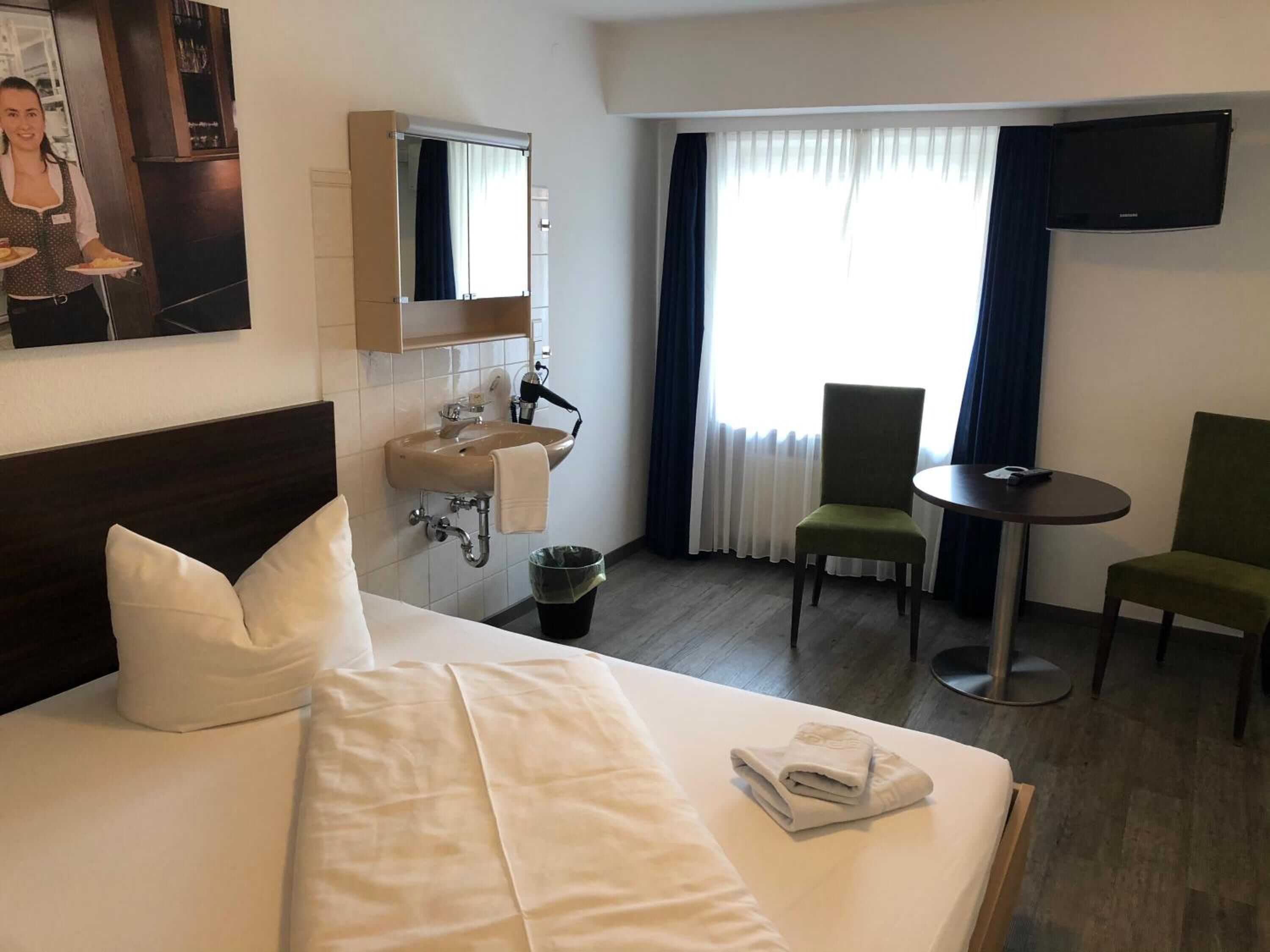 A hotel room with a bed, two chairs, a small table, a sink, and a mirror.