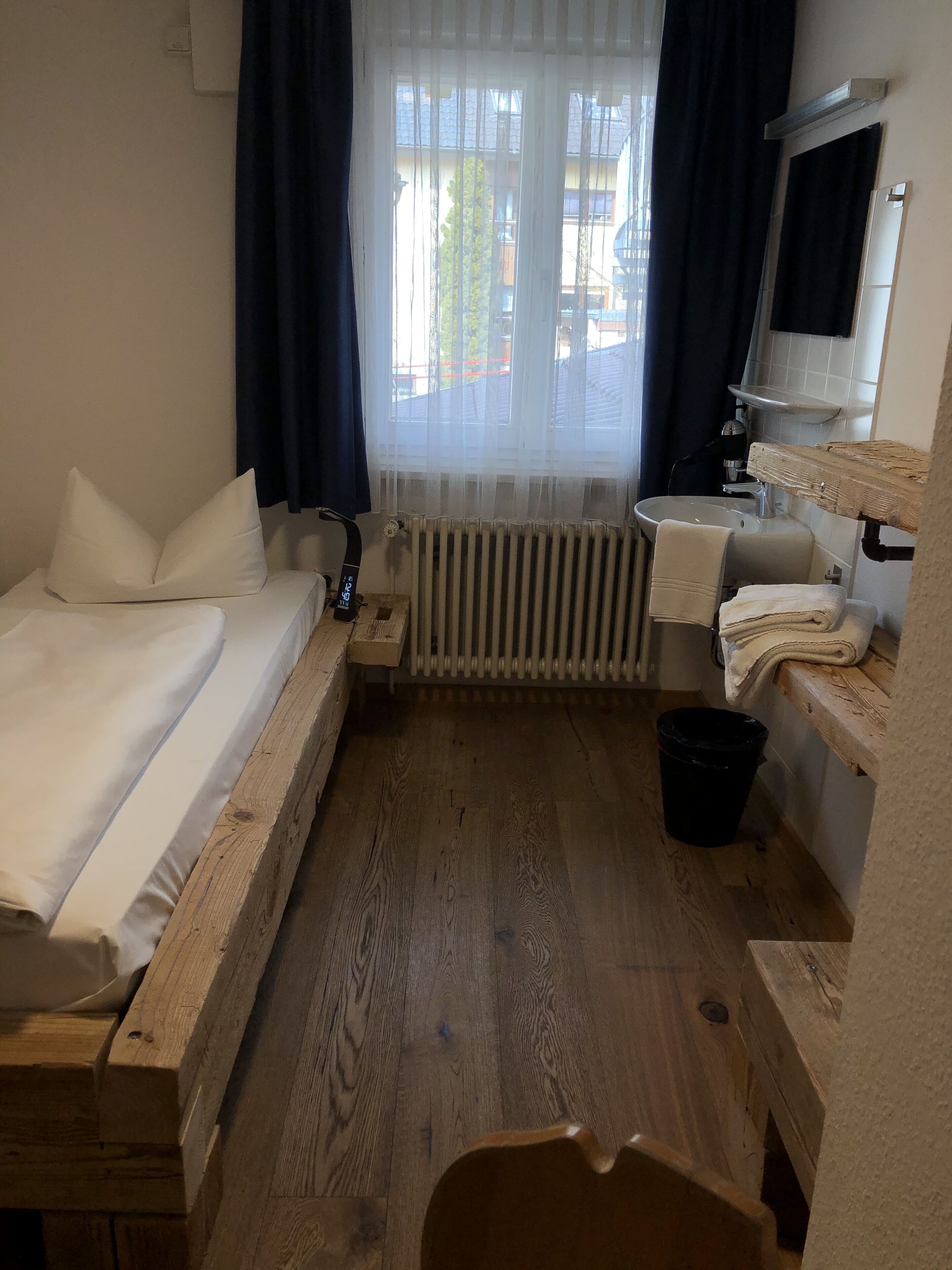 Quarto de hotel pequeno com cama de madeira, pia, espelho e vista para o exterior através de uma janela com cortinas leves.