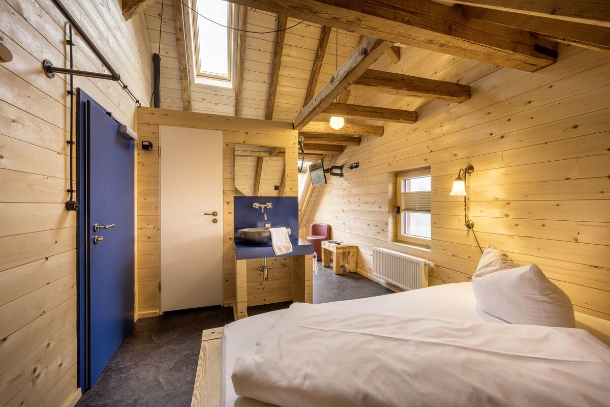 Een kamer met houten vloer, een bed, een blauwe deur, een wastafel en een raam.