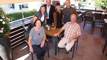 A group of people seated around a wooden table in an outdoor dining area with chairs and a potted plant.