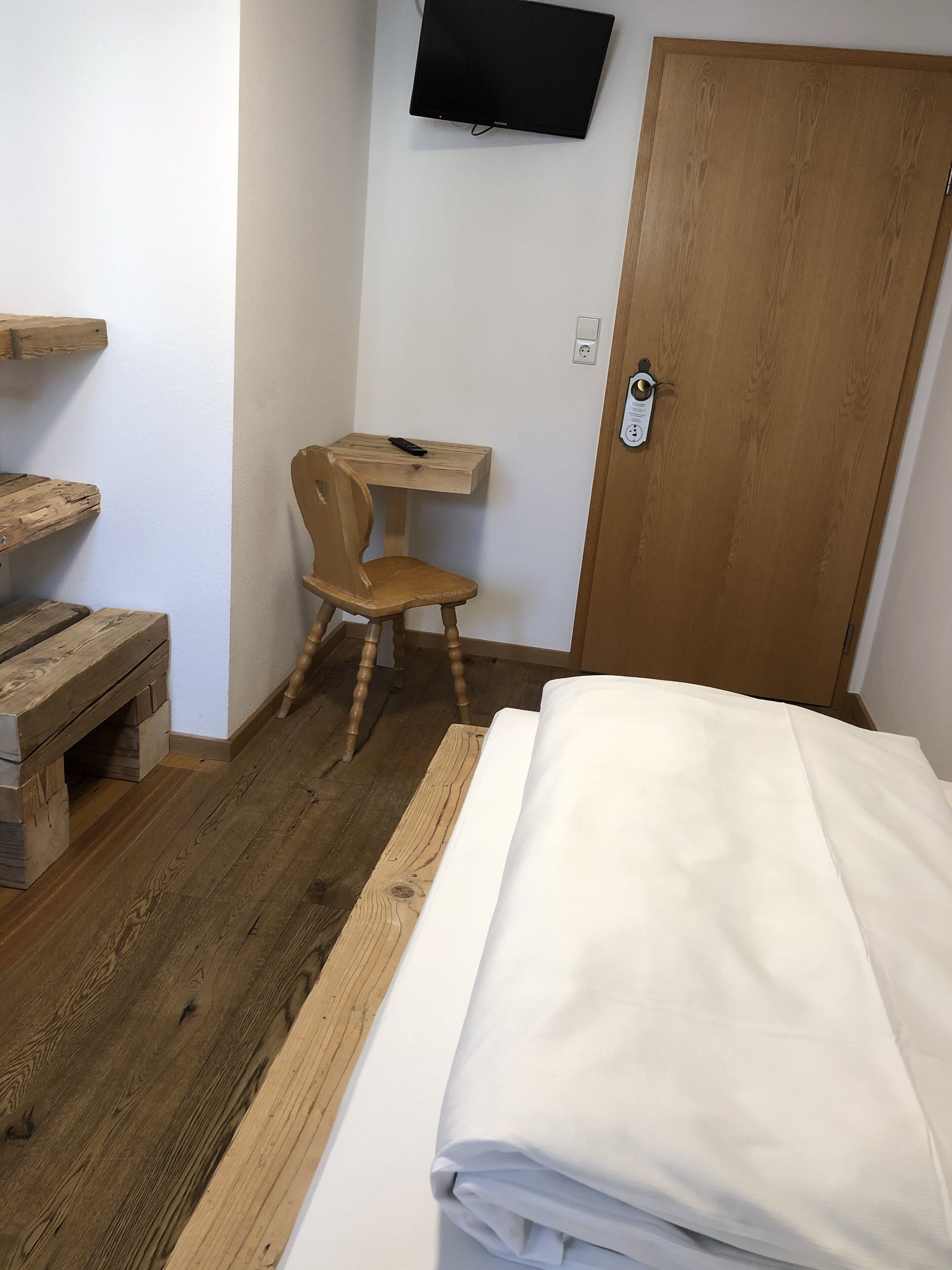 A bedroom with a wooden bed, a wooden desk with a chair, a television on the wall, and a wooden door.